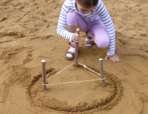 Ein Mädchen zeigt in einem Sandkasten, wie man eine Ellipse zeichnen kann, um beispielsweise ein ellipsenförmiges Gartenbeet anzulegen. (Foto: Hans-Joachim Vollrath)